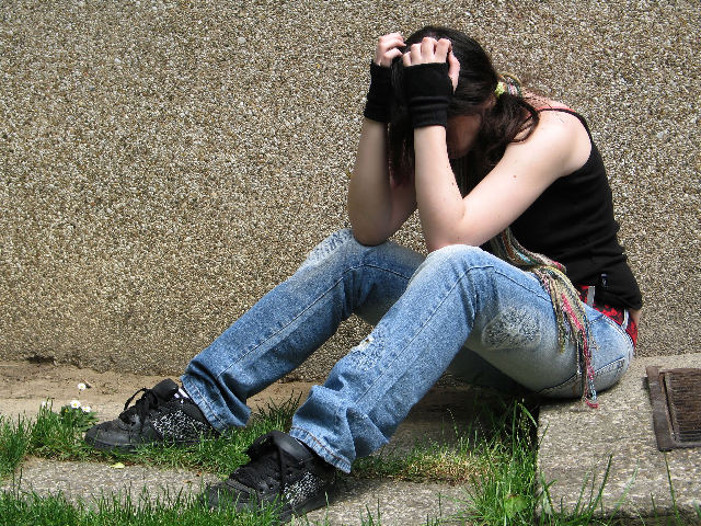 Young woman sits on the floor outside her flat, looking depressed